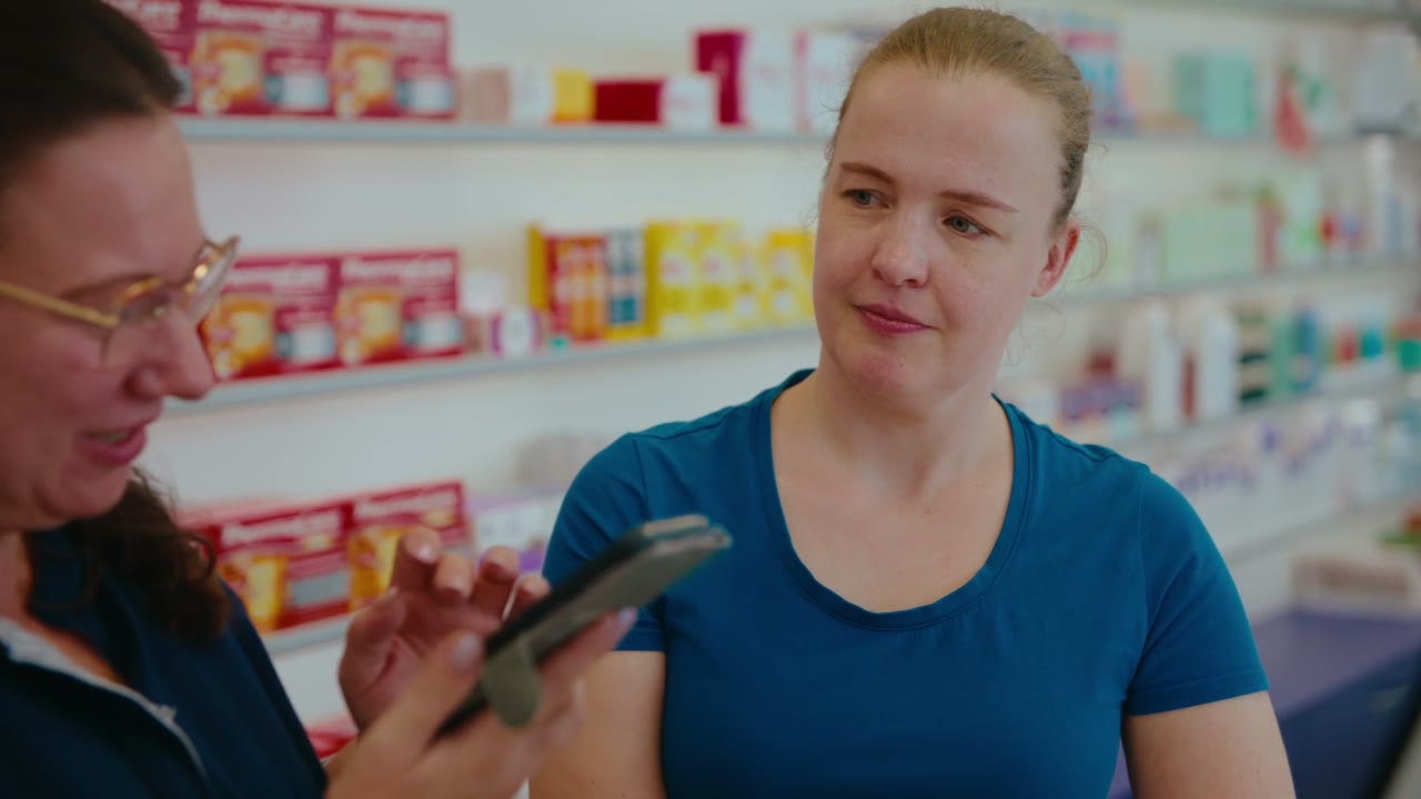 Szene aus dem Regionalfilm Börde: eine junge Frau in einer Apotheke, daneben eine Kollegin mit Smartphone