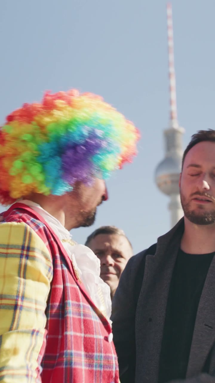 Szene aus dem Präventionsfilm im Hochformat: ein Mann mit bunter Clownsperücke, daneben sein Begleiter, hinten der Fernsehturm