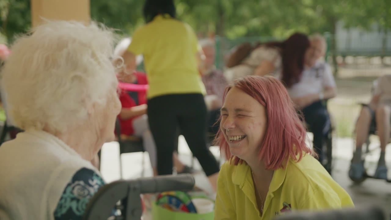 Eine junge Frau mit rosa Haaren lacht mit einer älteren Dame, Szene aus dem BMAS-Regionalfilm in Halle
