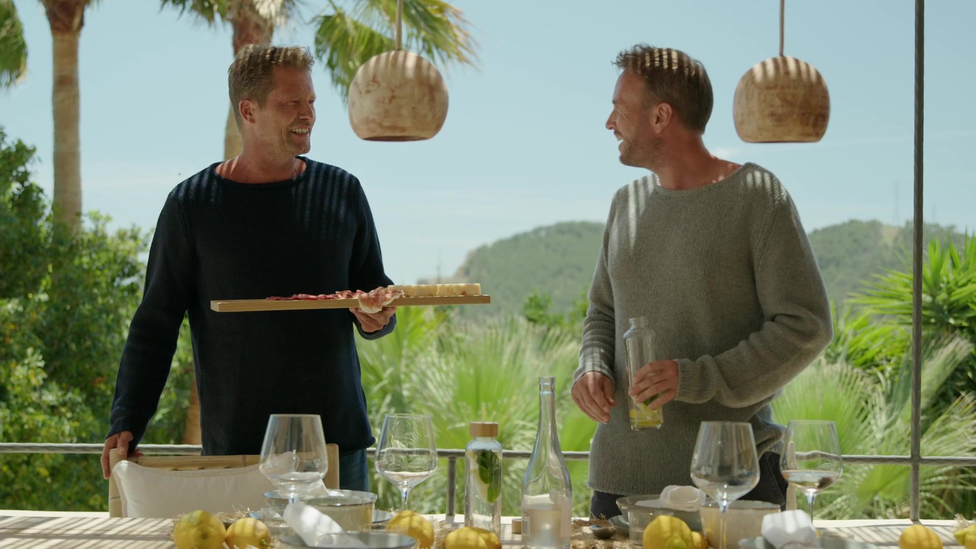 Two men laugh at a set table in a Mediterranean garden