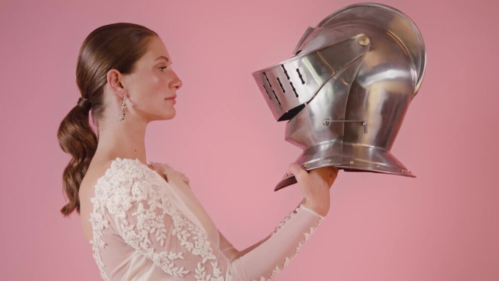 Bride in a lace dress holds a silver knight's helmet in profile