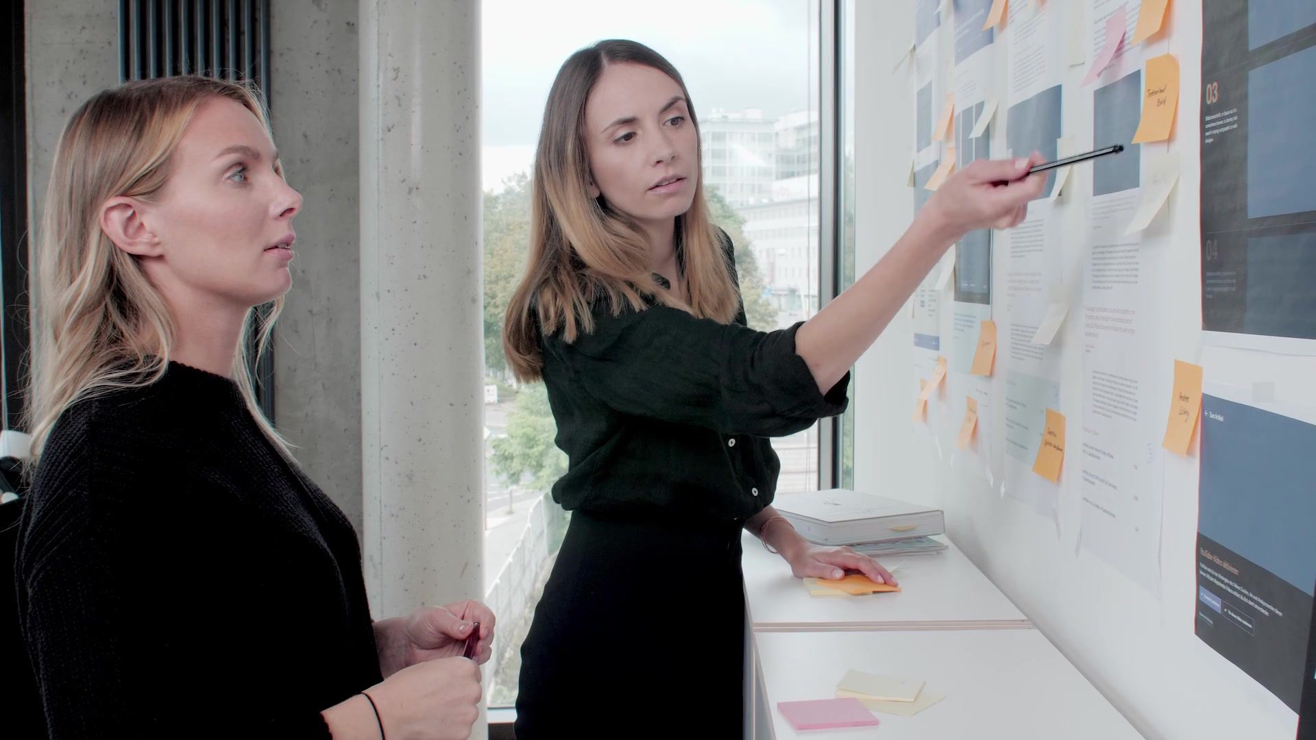 Two colleagues discuss wireframes and notes on the project wall