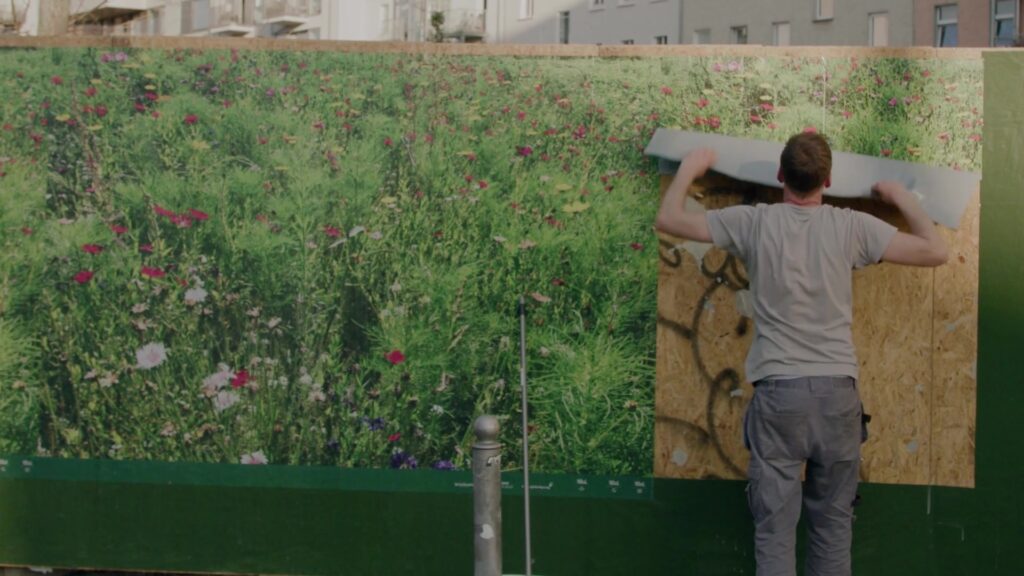 Mitarbeiter bringt großflächiges Blumenwiesen-Plakat an einer Bauzaunfläche an