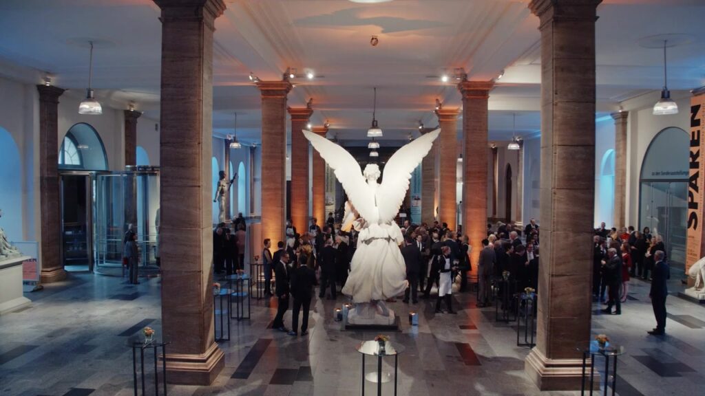 Festlich beleuchtete Museumshalle mit weißer Engelsskulptur und Gästen