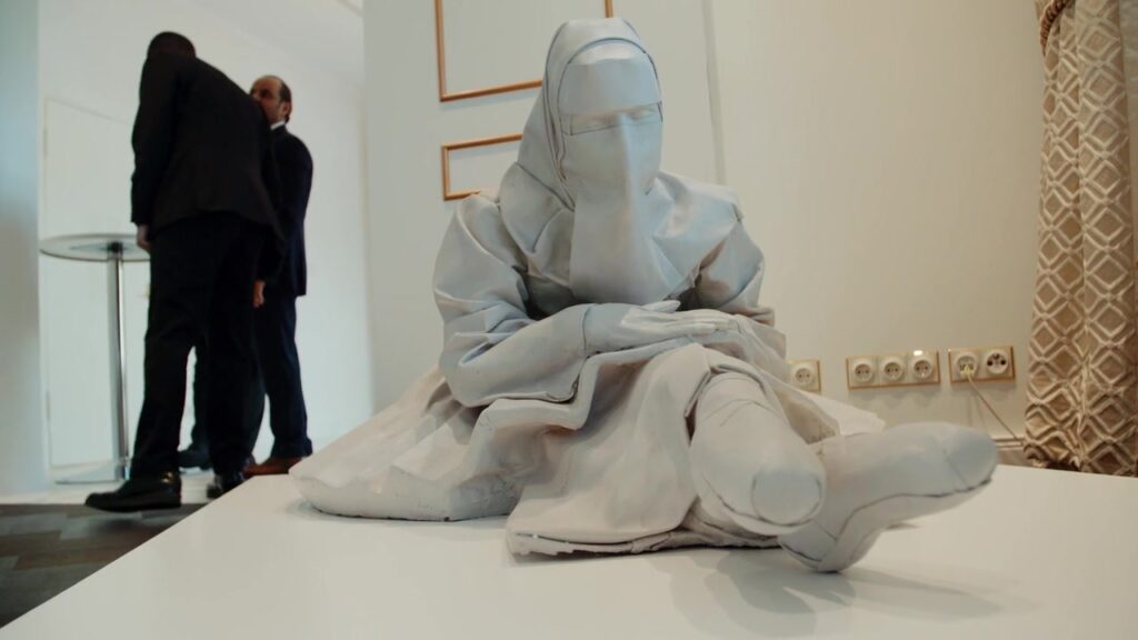 Weiße Skulptur einer sitzenden verschleierten Figur im Ausstellungsraum
