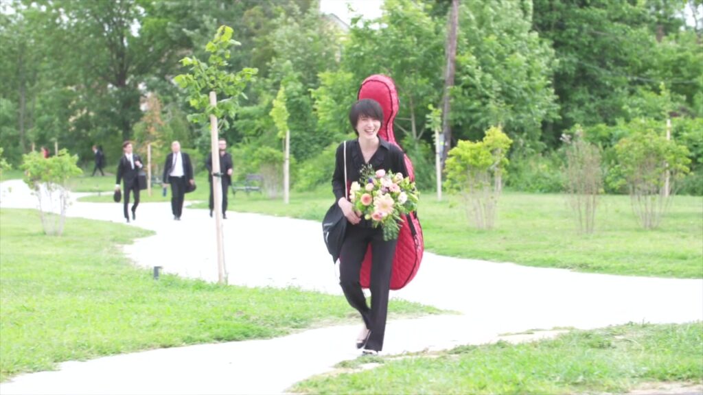 Lachende Musikerin mit rotem Cellokoffer und Blumenstrauß im Schlosspark